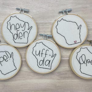 Peut inclure: Cinq pièces d'art en broderie sur cerceau représentant l'état du Wisconsin avec différentes phrases, dont "Hey Der", "Uff-da", "Ope!" et "Ope!"