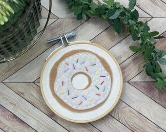 May include: A white and brown felt donut with colorful sprinkles, embroidered on a white felt background, displayed in a wooden embroidery hoop.