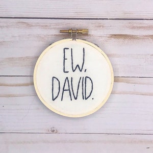 May include: A round embroidery hoop with the text "EW. DAVID." stitched in dark blue thread on white fabric. The hoop is made of light-colored wood and has a brass-colored tightening mechanism.