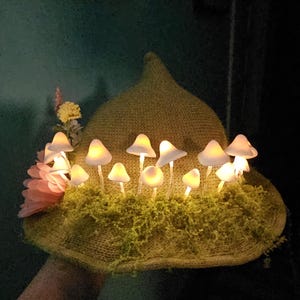 Pode incluir: Um chapéu verde claro adornado com luzes de cogumelos brilhantes e musgo artificial. Uma flor rosa e detalhes amarelos adicionam um toque de cor. Um acessório único e feito à mão.