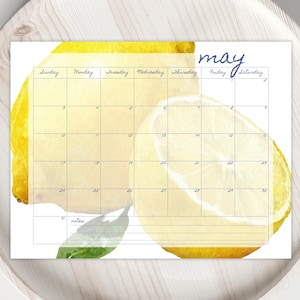 Könnte beinhalten: Ein Mai-Kalender mit einem Aquarell-Zitronen-Design. Der Kalender hat ein Rasterlayout mit Wochentagen und Daten. Das Wort "Mai" ist in Schreibschrift geschrieben. Der Kalender liegt auf einem hellen Holztablett.