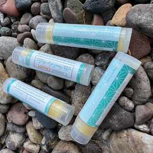May include: Four clear lip balm tubes with a teal and white geometric design. The tubes are labeled "The Best Lip Balm", "Coastal Desert", "Spearmint", and "0.15 oz". The tubes are sitting on a bed of small, gray rocks.