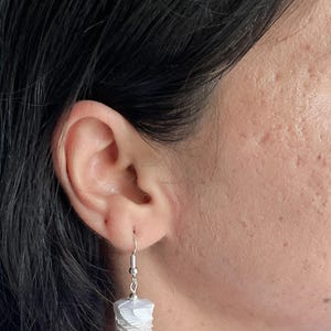 May include: A pair of dangle earrings with a white and purple layered design. The earrings are made of a woven material and have a silver hook.