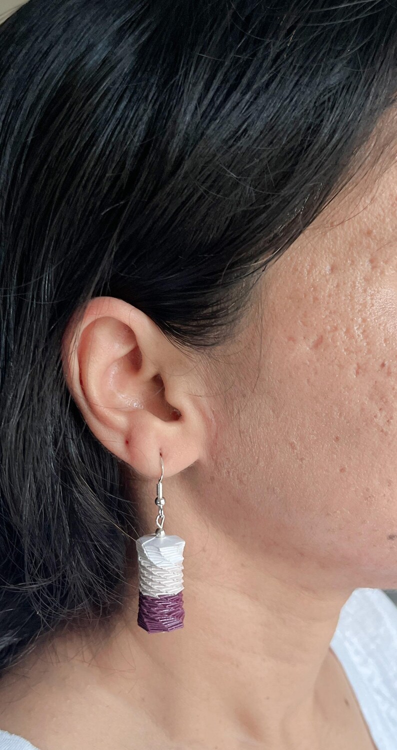 May include: A pair of dangle earrings with a white and purple layered design. The earrings are made of a woven material and have a silver hook.