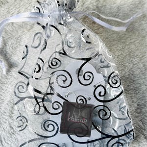 May include: A white organza bag with a black swirl pattern. The bag contains a pair of silver hoop earrings and a small white card with the word "Kalaap" printed on it.