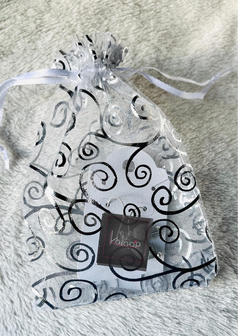 May include: A white organza bag with a black swirl pattern. The bag contains a pair of silver hoop earrings and a small white card with the word "Kalaap" printed on it.