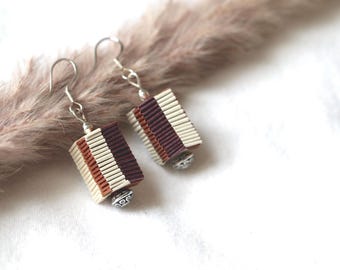 May include: A pair of earrings with rectangular shaped pendants. The pendants are made of woven fabric in shades of brown, white, and purple. The earrings have silver hooks and silver bead accents.
