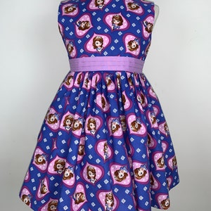 May include: A purple and blue sleeveless dress with a heart pattern featuring a cartoon princess. The dress has a purple sash around the waist.