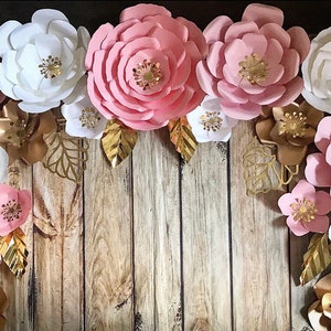 May include: A collection of large paper flowers in pink and white with gold accents. The flowers are arranged on a wood background.
