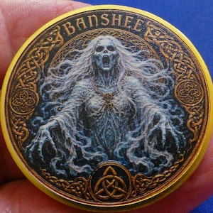 May include: A circular gold-toned metal coin featuring a detailed illustration of a banshee with flowing white hair and outstretched hands. The word BANSHEE is printed at the top, and a triquetra knot symbol is centered at the bottom edge.