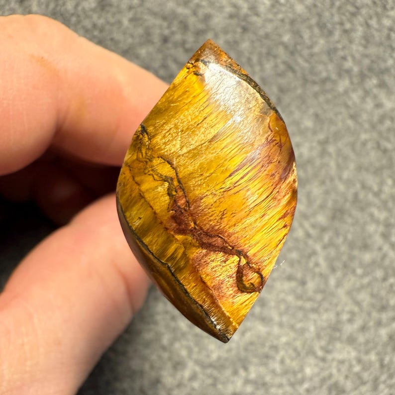 Puede incluir: Una piedra preciosa pulida en forma de l&aacute;grima, de color amarillo dorado y con intrincadas vetas marrones. La piedra se sostiene en una mano, mostrando sus patrones y texturas naturales. La superficie de la piedra preciosa refleja la luz.
