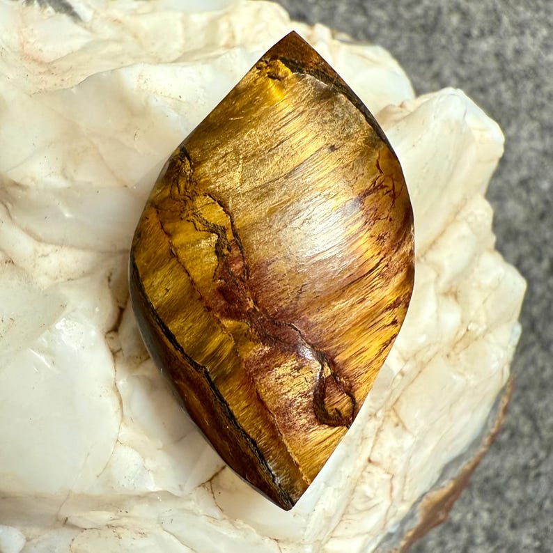 Puede incluir: Una piedra preciosa de ojo de tigre pulida en forma de l&aacute;grima. La piedra presenta una mezcla de tonos amarillo dorado y marr&oacute;n con estr&iacute;as naturales. La piedra preciosa est&aacute; sobre una superficie rocosa blanca y beige.