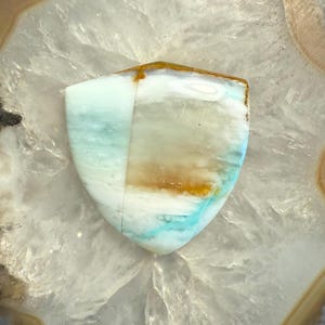 May include: A light blue, white, and brown agate cabochon with a shield shape. The stone has a natural, organic pattern.