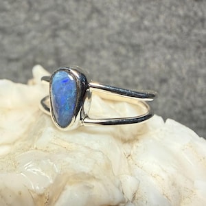 May include: A silver ring featuring a pear-shaped, blue opal gemstone. The opal has a vibrant, iridescent quality. The ring's band has a split design, with two parallel silver bands. The ring is set against a neutral background.