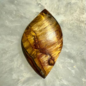 Puede incluir: Una piedra preciosa de ojo de tigre pulida en forma de l&aacute;grima. La piedra muestra una rica mezcla de tonos amarillo dorado y marr&oacute;n, con chatoyancy natural que crea una apariencia brillante y sedosa. La piedra preciosa est&aacute; cortada y pulida, mostrando sus patrones &uacute;nicos.