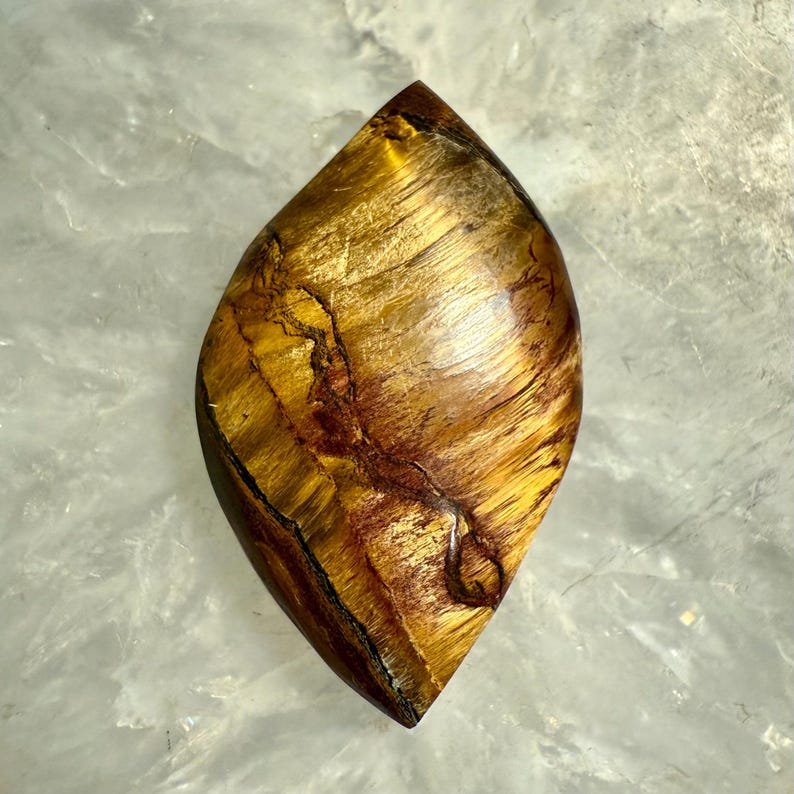 Puede incluir: Una piedra preciosa de ojo de tigre pulida en forma de l&aacute;grima. La piedra muestra una rica mezcla de tonos amarillo dorado y marr&oacute;n, con chatoyancy natural que crea una apariencia brillante y sedosa. La piedra preciosa est&aacute; cortada y pulida, mostrando sus patrones &uacute;nicos.