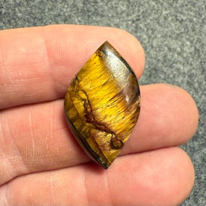 Puede incluir: Una piedra preciosa de ojo de tigre pulida en forma de l&aacute;grima. La piedra presenta bandas de tonos amarillo dorado y marr&oacute;n, con un efecto chatoyant natural. La gema se sostiene en una mano, mostrando sus patrones y brillo &uacute;nicos.