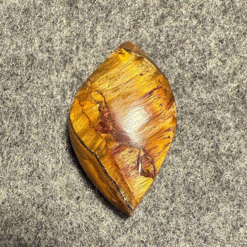 Puede incluir: Una piedra preciosa de ojo de tigre pulida en forma de l&aacute;grima. La piedra presenta una mezcla de tonos amarillo dorado y marr&oacute;n con patrones naturales y un acabado brillante. La piedra preciosa est&aacute; sobre un fondo gris.