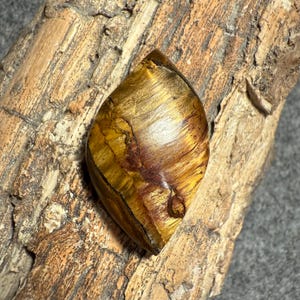 Puede incluir: Una gema pulida en forma de l&aacute;grima con un patr&oacute;n arremolinado de tonos dorados, marrones y &aacute;mbar. La piedra se encuentra sobre un fondo de madera natural texturizada, resaltando sus variaciones de color &uacute;nicas.