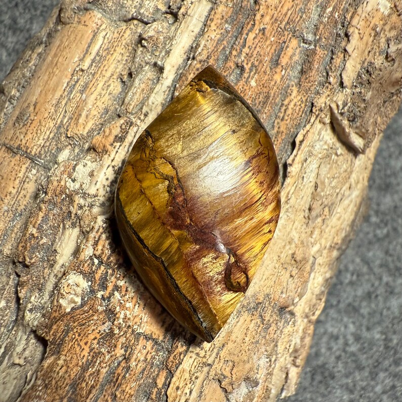 Puede incluir: Una gema pulida en forma de l&aacute;grima con un patr&oacute;n arremolinado de tonos dorados, marrones y &aacute;mbar. La piedra se encuentra sobre un fondo de madera natural texturizada, resaltando sus variaciones de color &uacute;nicas.