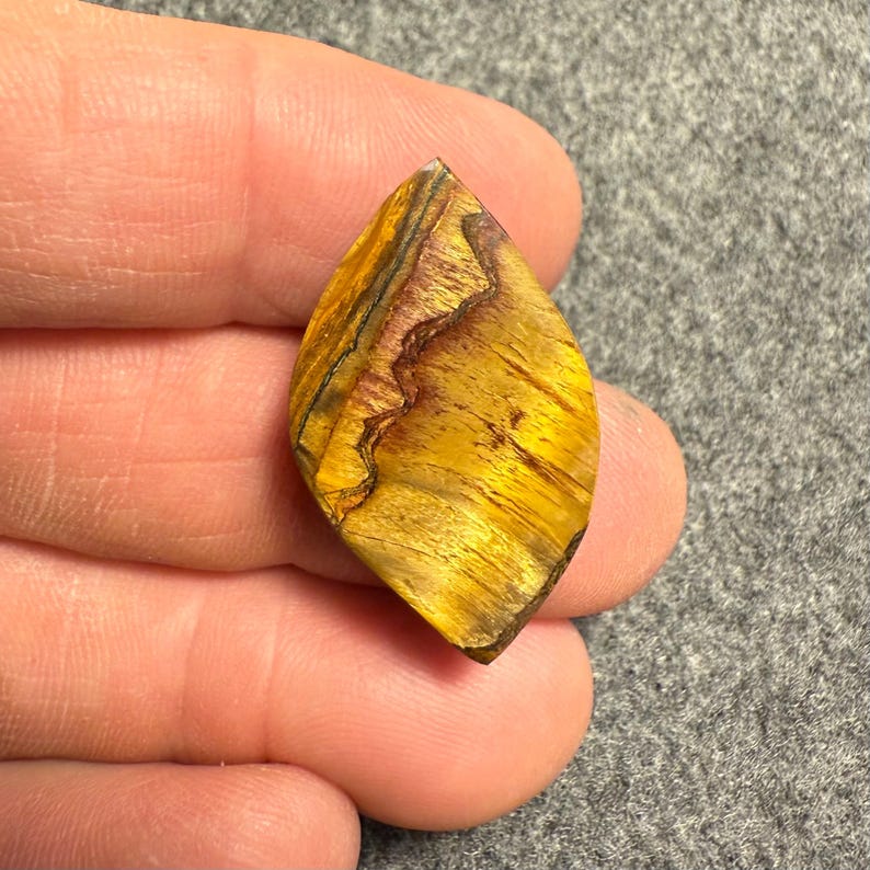 Puede incluir: Una gema pulida en forma de l&aacute;grima con un llamativo patr&oacute;n de tonos amarillo dorado, marr&oacute;n y negro. La superficie de la piedra muestra una veta natural &uacute;nica, creando una textura visualmente atractiva. La gema se sostiene en una mano.