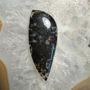 May include: A black and white agate cabochon with a unique pattern and a hint of blue. The cabochon is shaped like a teardrop and has a smooth, polished surface.