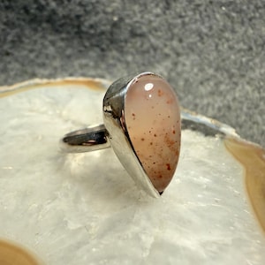 May include: A silver ring featuring a teardrop-shaped, light pink gemstone with brown speckles. The gemstone is set in a polished silver bezel, and the ring band is also silver.