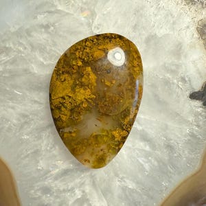 May include: A smooth, polished agate stone with a unique pattern of brown and gold dendrites. The stone has a natural, organic shape and a translucent appearance.