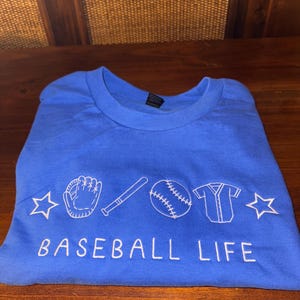 May include: A royal blue t-shirt with a white embroidered design of a baseball glove, bat, ball, and jersey. The text "BASEBALL LIFE" is also embroidered in white.