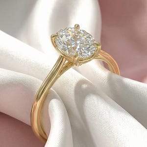 Könnte beinhalten: Ein goldener Verlobungsring mit einem großen, klaren Diamanten im Kissenschliff. Der Ring hat ein zartes Goldband und liegt auf weißem und rosa Satin.