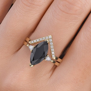 Könnte beinhalten: Ein goldener Ring mit einem schwarzen, markisenförmigen Mittelstück und kleineren Diamant-Akzenten. Der Ring ist auf einem V-förmigen Band mit einer Reihe von Diamanten gesetzt.