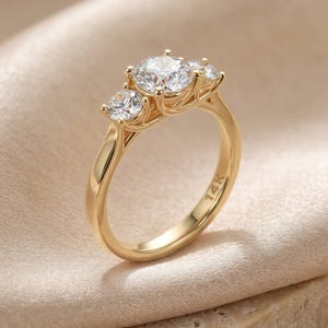 May include: A gold ring featuring three round clear stones set in a prong setting. The central stone is larger than the two flanking stones. The inner band of the ring is clearly engraved with the text 14K, indicating the gold purity of the metal.