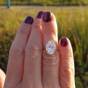 Oval moissanite engagement ring displayed on a hand outdoors, with a prong-set center stone and pavé shoulders.