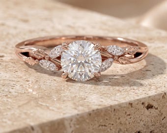 Può includere: Un anello in oro rosa con una grande pietra centrale rotonda a taglio brillante. La fascia è decorata con delicati dettagli metallici a forma di foglia e piccole pietre a taglio marquise che fiancheggiano il diamante centrale.