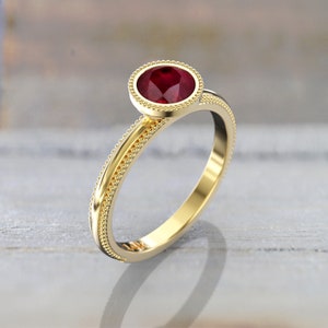 Three-quarter view of a yellow gold engagement ring featuring a faceted round red ruby, a smooth bezel with beaded edging, and decorative beading along the band.