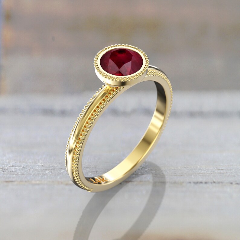 Three-quarter view of a yellow gold engagement ring featuring a faceted round red ruby, a smooth bezel with beaded edging, and decorative beading along the band.