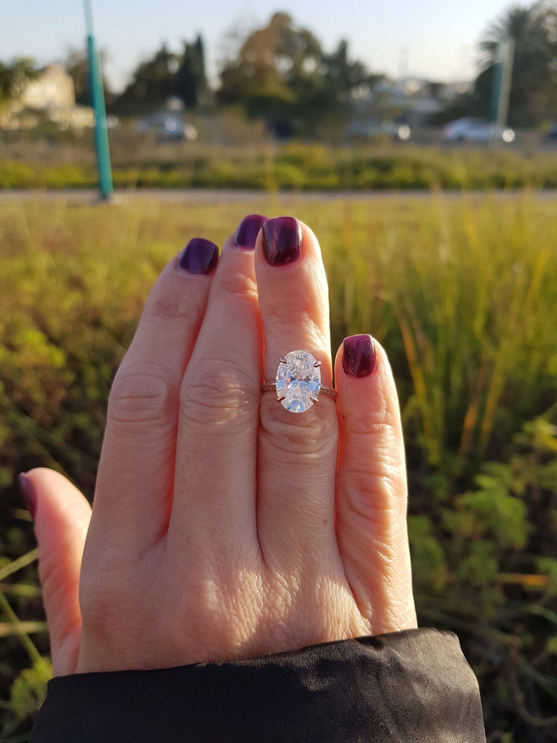 Oval moissanite engagement ring displayed on a hand outdoors, with a prong-set center stone and pavé shoulders.
