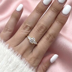 May include: A gold ring with a large, round center stone and two smaller pear-shaped stones on either side. The ring is set on a hand with white nail polish, against a pink satin background.