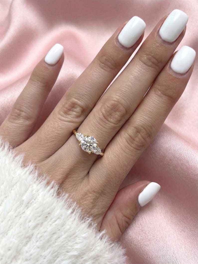 May include: A gold ring with a large, round center stone and two smaller pear-shaped stones on either side. The ring is set on a hand with white nail polish, against a pink satin background.