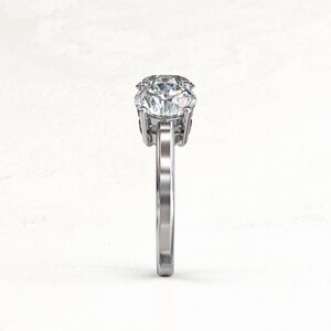 Side-on structural view of a white/silver-colored three-stone moissanite ring, highlighting the raised center setting, prongs, open gallery, and narrow polished band.