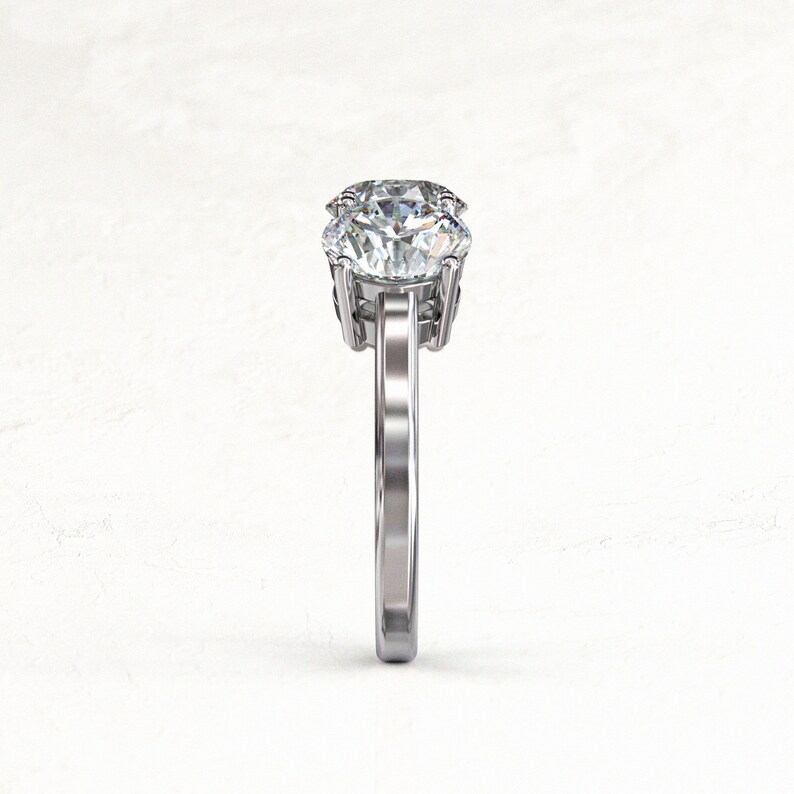 Side-on structural view of a white/silver-colored three-stone moissanite ring, highlighting the raised center setting, prongs, open gallery, and narrow polished band.