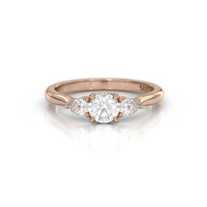 May include: Rose gold ring featuring a large, round, clear gemstone flanked by two smaller, pear-shaped gemstones. The band is smooth and polished, with a subtle shine. The ring is set against a white background.