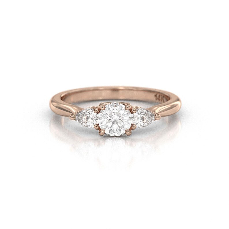 May include: Rose gold ring featuring a large, round, clear gemstone flanked by two smaller, pear-shaped gemstones. The band is smooth and polished, with a subtle shine. The ring is set against a white background.