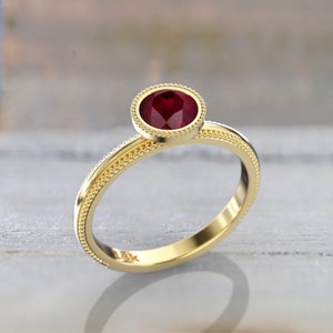 Angled close-up of a yellow gold round ruby engagement ring with a smooth bezel, beaded stone border, and matching beaded detailing along the band.