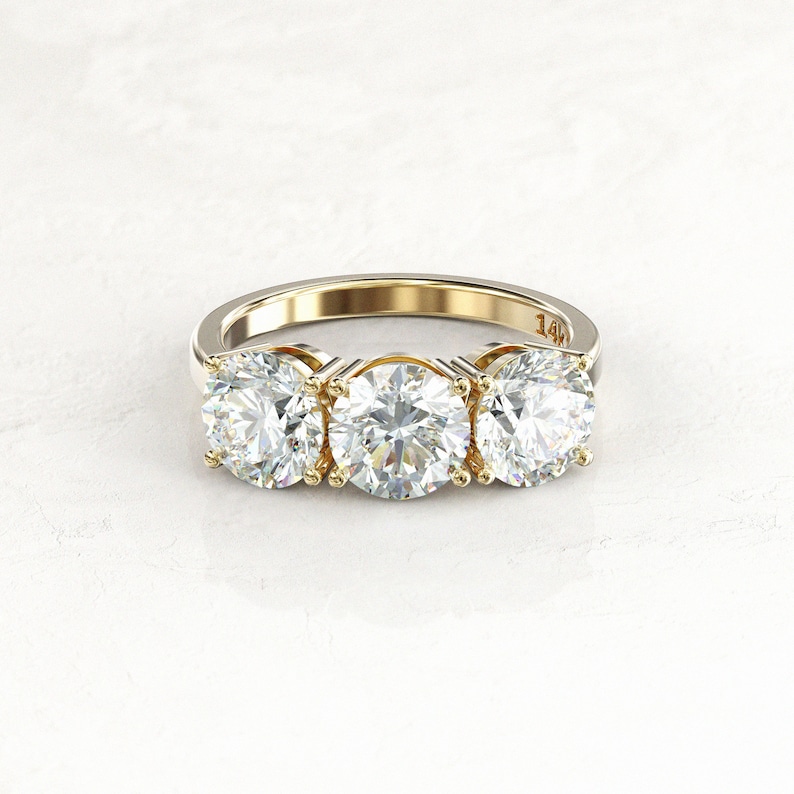 Close low-angle front view of a two-tone three-stone moissanite engagement ring, showing yellow gold prongs and inner settings, a white/silver-colored band, and a 14K stamp.