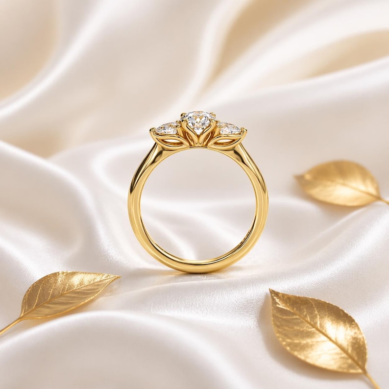 May include: A gold ring with a three-stone design, featuring a large central diamond and two smaller diamonds on either side. The ring is set against a backdrop of white satin fabric and gold leaf accents.