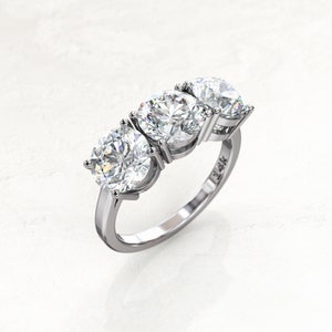 Angled overhead view of a white/silver-colored three-stone moissanite engagement ring, showing the larger round center stone, two side stones, raised prongs, open settings, and polished band.