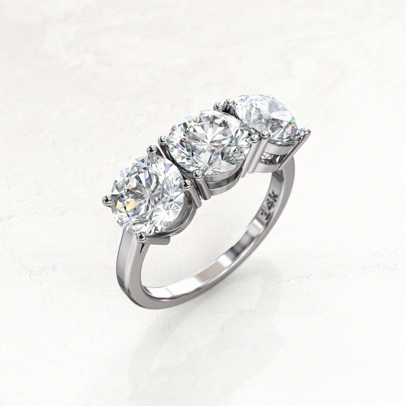Angled overhead view of a white/silver-colored three-stone moissanite engagement ring, showing the larger round center stone, two side stones, raised prongs, open settings, and polished band.