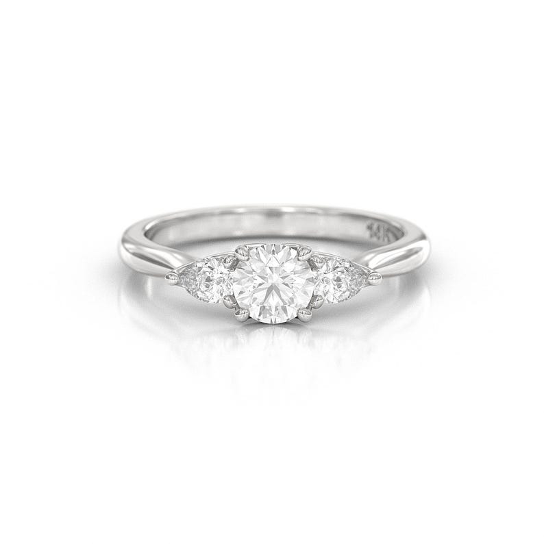 May include: A silver ring with a large, round, clear gemstone in the center, flanked by two smaller, pear-shaped gemstones. The ring band is smooth and polished.
