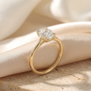 Könnte beinhalten: Ein goldener Ring mit einem einzelnen, oval geschliffenen, klaren Edelstein in einer Vier-Krappen-Fassung. Die Ringschiene ist glatt und poliert und zeigt ein klassisches Solitär-Design mit einem schlanken Profil, das den Glanz des Steins betont.
